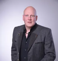 Ein kahlköpfiger Mann in einem grauen Blazer mit schwarzem Hemd und Halskette posiert für ein Porträt.
