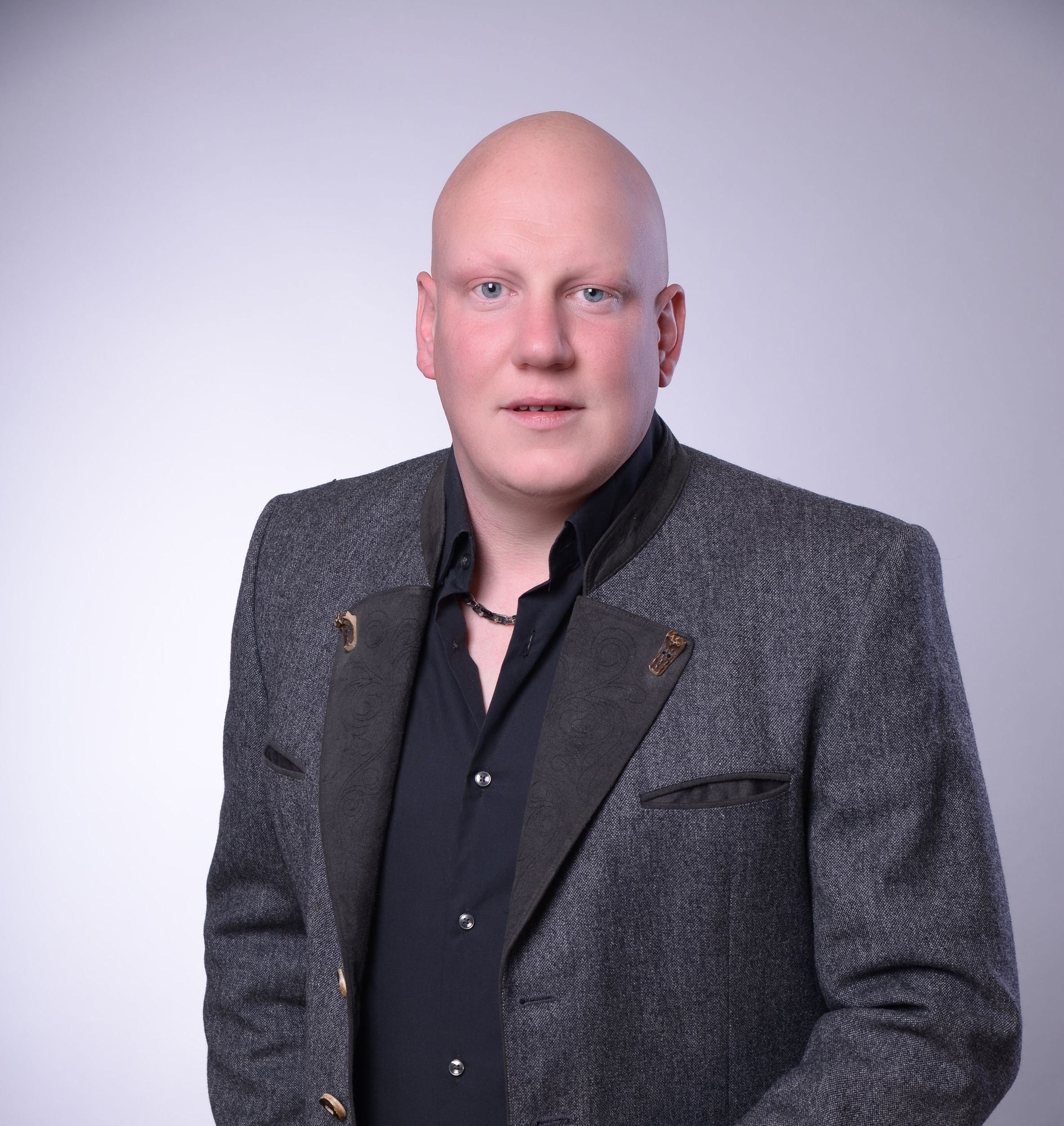 Ein kahlköpfiger Mann in einem grauen Blazer mit schwarzem Hemd und Halskette posiert für ein Porträt.