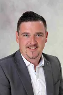 Ein Mann mit kurzem Haarschnitt, der einen grauen Blazer und ein weißes Hemd trägt, lächelt und scheint für ein Foto zu posieren.