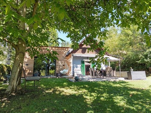Ein modernes Haus mit Holzverkleidung und einem Terrassenbereich mit Tisch und Stühlen unter einem Regenschirm. Ein Hängesessel schwingt neben einem Baum.