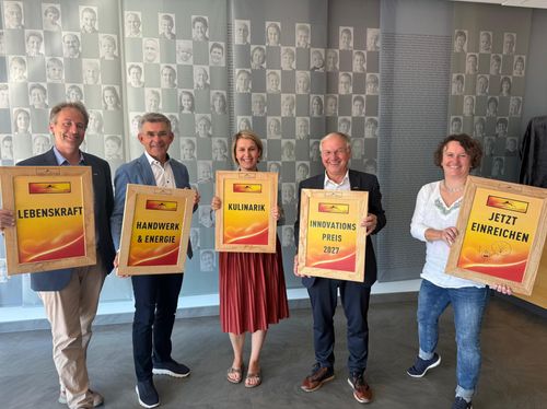 Fünf Menschen stehen zusammen und halten gerahmte Urkunden. Der Mann auf der linken Seite hält eine Urkunde mit dem Titel 'Handwerk & Energie'. Die Frau in der Mitte hält eine Urkunde mit dem Titel 'KulinariK'. Der Mann auf der rechten Seite hält eine Urkunde mit dem Titel 'Innovations Preis 2027'. Die Frau ganz rechts hält eine Urkunde mit dem Titel 'E'. Hinter ihnen ist eine Wand mit vielen kleinen Fotos von Menschen.