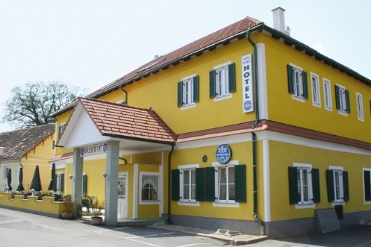 Ein gelbes Gebäude mit rotem Dach und grünen Fensterläden, als Hotel beschriftet, steht in einem ländlichen Gebiet mit Bäumen und geschlossenen Schirmen.