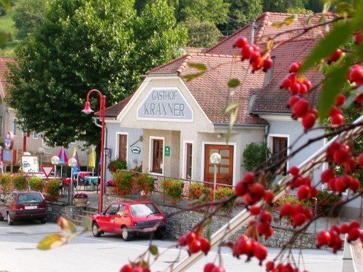 Das Gasthof Kraxner Gebäude mit rotem Dach hat ein Schild über dem Eingang. Rote Beeren hängen von Bäumen davor. Autos sind auf der Seite geparkt.
