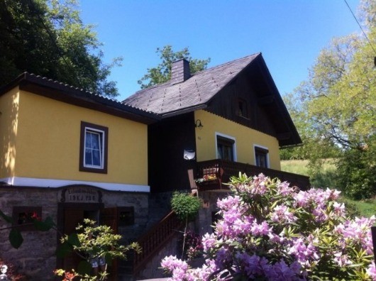 Ein gelbes und schwarzes Haus mit einem Schornstein, umgeben von Bäumen und blühenden Pflanzen. Es hat eine Holztreppe und einen Balkon mit Geländer.