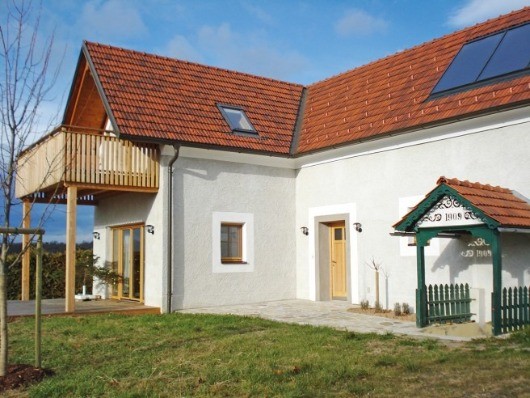 Ein weißes Haus mit rotem Dach und Solarpanelen, mit Balkon, Holztüren und grünen Zäunen im Garten.