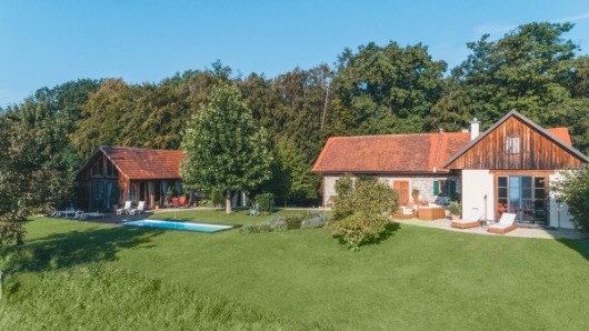 Zwei Häuser mit roten Dächern befinden sich in einer grünen Landschaft. Eines hat einen Swimmingpool. Bäume und Pflanzen umgeben das Grundstück.