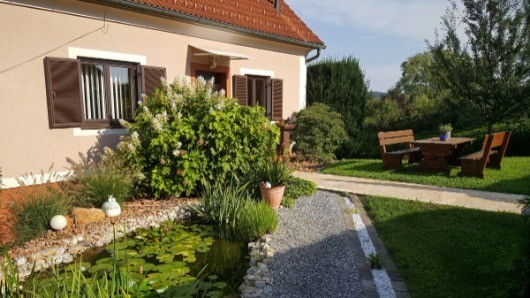 Ein Haus mit einem braunen Dach, einem Holztisch und Bänken, einem Teich mit Seerosen und einem Garten mit Pflanzen und Sträuchern.