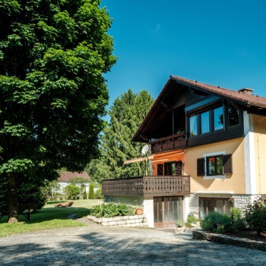 Ein gelbes und weißes Haus mit braunem Dach und Balkon, umgeben von Bäumen und Pflanzen, mit einer Einfahrt vor dem Haus.