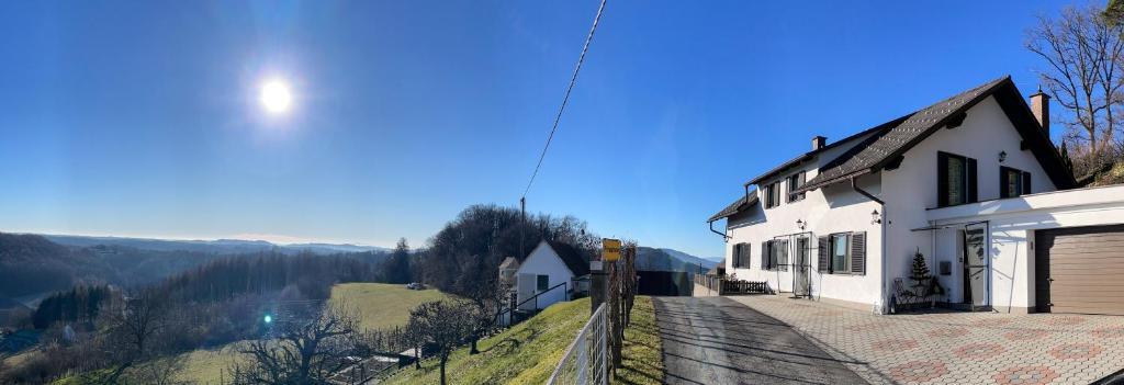 Ein sonniger Tag zeigt ein Haus mit einer Einfahrt, einem Zaun und einem kleineren Haus in der Nähe. Die Landschaft umfasst Bäume, Berge und einen klaren blauen Himmel.