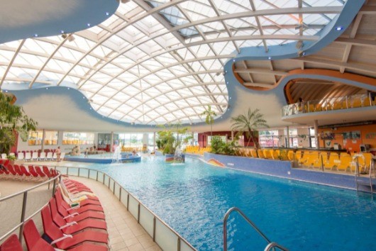 Ein Hallenbad mit Glasdach und blauen Wänden. Rote Liegestühle und gelbe Stühle stehen um den Pool herum. Es gibt Topfpflanzen und eine Bartheke.