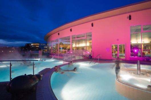 Ein Außenpool mit rosa Wänden und Fenstern ist bei Nacht beleuchtet. Es gibt Menschen, die im Pool schwimmen.
