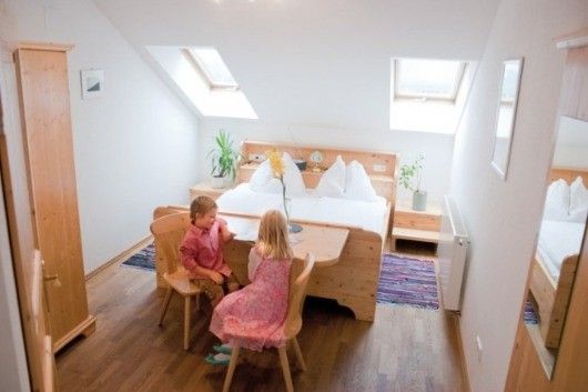 Zwei Kinder sitzen an einem Schreibtisch in einem Raum mit Holzboden, einem Bett mit weißen Laken und einem Spiegel. In der Nähe stehen Topfpflanzen und ein Holzschrank.