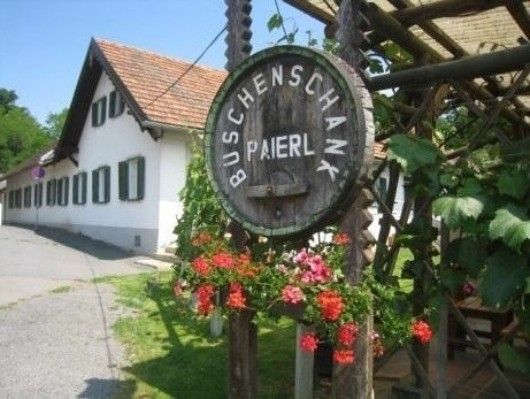Ein großes hölzerne Fassschild für Buschenschank Paierl hängt vor einem weißen Gebäude mit rotem Dach. Das Schild ist von Blumen und Grün umgeben.