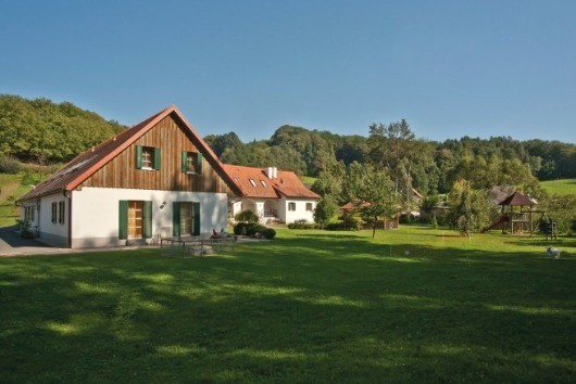 Eine ländliche Landschaft mit einem weißen Haus mit brauner Einfassung und grünen Fenstern und einem braunen Dach im Vordergrund. Im Hintergrund befinden sich weitere Häuser, Bäume und ein klarer blauer Himmel.
