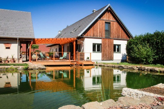 Ein Holzhaus mit einem Teich davor. Das Haus hat eine Holzterrasse mit Stühlen und einem kleinen Baum.