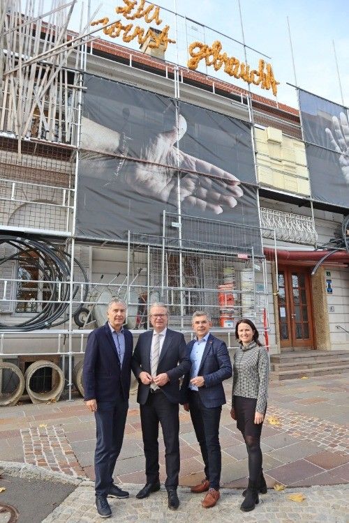Vier Personen in formeller Kleidung stehen vor einem Gebäude im Bau, mit Gerüsten und einem großen Banner mit Händen.