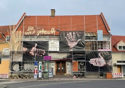 Ein Gebäude in Renovierung mit rotem Dach und Gerüsten, das zwei große Handbilder auf Bannern und zusätzliche Baumaterialien auf der Straße zeigt.