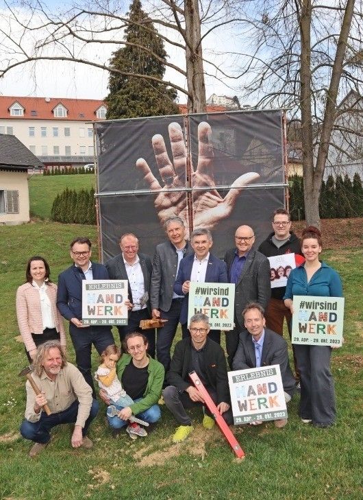 Gruppenfoto von Menschen vor einem Gebäude. Einige halten Schilder mit 'Wir sind Hand und Werk' und Datumsangaben. Ein Banner mit Händen ist im Hintergrund.
