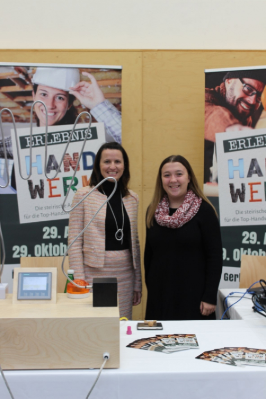 Zwei Frauen stehen neben einem Tisch mit Werbematerialien und lächeln. Hinter ihnen sind Plakate mit 'Handwerk' und einem Datum, 29. Oktober. Der Tisch hat einen Monitor, einen Lautsprecher und Flyer.