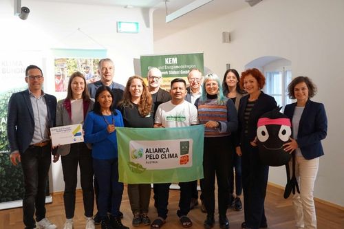 Eine Gruppe von Menschen steht in einem Raum und hält ein grünes Banner mit der Aufschrift 'Alianca Pelo Clima Austria.' Sie alle lächeln und scheinen für ein Foto zu posieren. Hinter ihnen ist ein grünes Banner mit Text zu sehen.