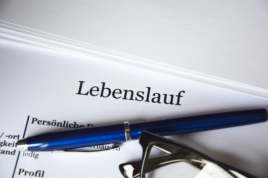 Ein Dokument mit dem Wort Lebenslauf liegt auf einem Schreibtisch. Ein blauer Stift und eine Brille liegen auf dem Dokument.