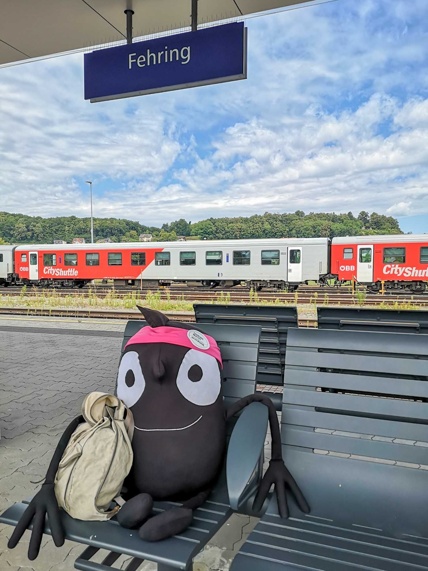 Ein CityShuttle-Zug steht auf einem Bahnhof. Ein großer Plüschtier sitzt auf einer Bank mit einem Rucksack. Der Bahnhof ist von Grün umgeben.