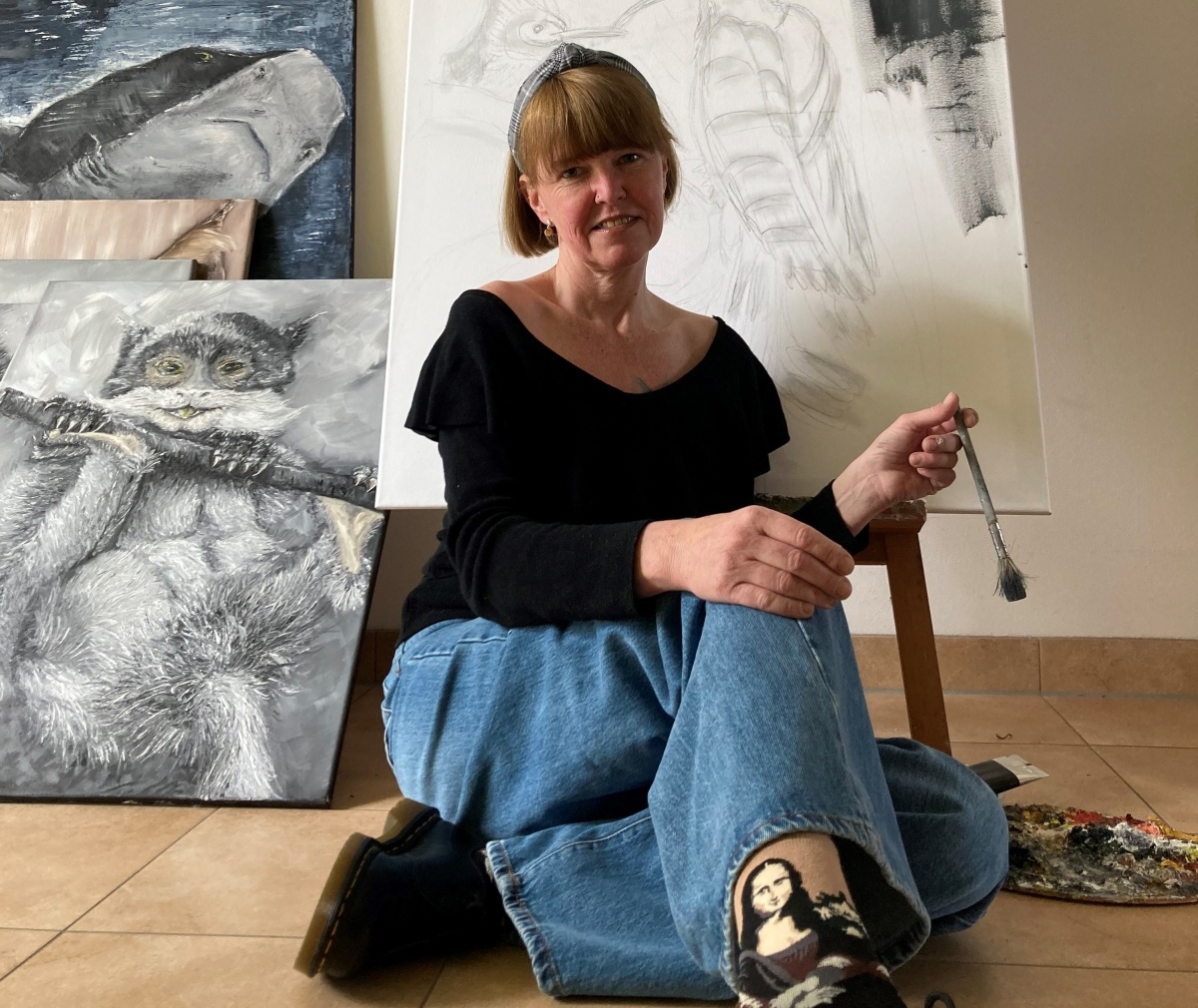 Eine Frau sitzt vor einer großen Leinwand auf dem Boden und hält einen Pinsel in der Hand. Sie trägt ein schwarzes Oberteil und Jeans. An der Wand hinter ihr hängen verschiedene Gemälde, darunter ein Hai und eine Eule.