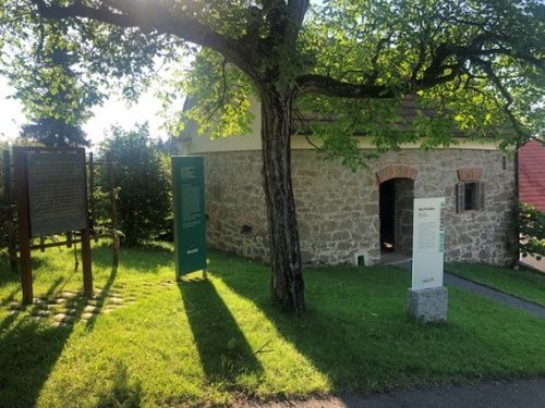 Ein kleines Steingebäude mit einem gewölbten Eingang, umgeben von einem Rasen. Zwei grüne Schilder stehen vor dem Gebäude.