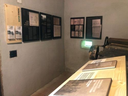 Ein kleiner Raum mit einem Holztisch, Büchern und historischen Dokumenten. Die Wand ist mit gerahmten Bildern und Texten versehen. Eine Maschine mit grünem Licht steht rechts.