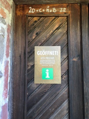 Eine Holztür mit einem Schild, auf dem 'GEOFFNET! ALTE PRESSHAUS' steht, einem 'i'-Symbol und zusätzlichem Text darunter. Die Tür hat die Nummer '20' und weiteren Text darüber.