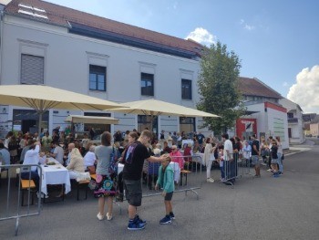 Eine Menschenmenge steht vor einem weißen Gebäude mit mehreren großen Sonnenschirmen. Sie stehen um Tische mit Essen herum und warten möglicherweise in einer Schlange. Vor der Menge befinden sich mehrere Metallbarrieren.