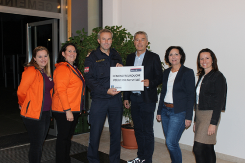 Fünf Menschen stehen lächelnd und halten eine Urkunde vor einem Gebäude. Drei Frauen und zwei Männer, von denen einer in einer Polizeiuniform ist.