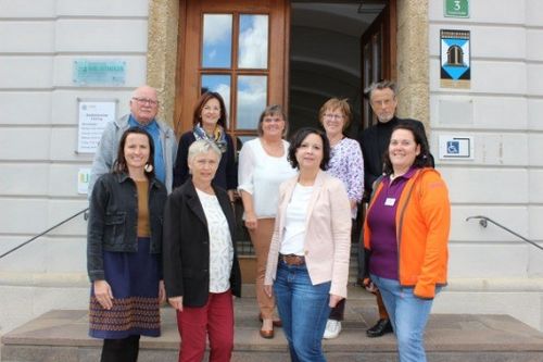 Eine Gruppe von Menschen, sowohl Männer als auch Frauen, stehen auf den Stufen eines Gebäudes und lächeln für ein Foto. Hinter ihnen befindet sich eine Holztür mit Glasfenstern.