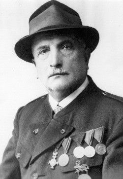 Eine Schwarz-Weiß-Porträt eines Mannes in Militäruniform. Er trägt einen Fedora und mehrere Medaillen auf der Brust.