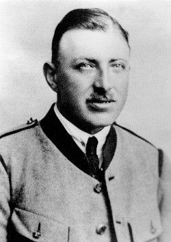Ein schwarz-weißes Bild eines Mannes in Uniform mit ernster Miene, geschmückt mit einer Medaille und Knöpfen an seiner Jacke.