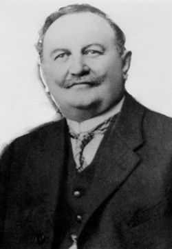 Ein schwarz-weißes Porträt eines Mannes in Anzug und Krawatte mit einem Schnurrbart, der nach vorne schaut.