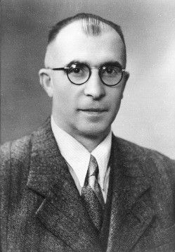 Schwarz-Weiß-Porträt eines Mannes in formeller Kleidung, der eine Brille und eine Krawatte trägt, direkt in die Kamera mit einem ernsten Ausdruck schaut.