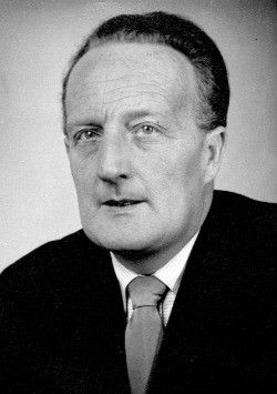 Ein Schwarz-Weiß-Foto eines Mannes in Anzug und Krawatte, der mit ernster Miene direkt in die Kamera schaut.
