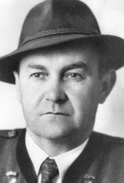 Schwarz-Weiß-Foto eines Mannes in Uniform, trägt einen Hut mit Abzeichen, eine Krawatte und ein militärisches Abzeichen auf seiner linken Schulter. Er hat einen ernsten Gesichtsausdruck.