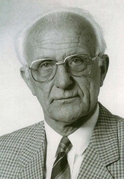 Ein älterer Mann mit weißen Haaren, der einen karierten Anzug, ein weißes Hemd und eine gestreifte Krawatte trägt. Er trägt eine Brille und hat einen ernsten Gesichtsausdruck.