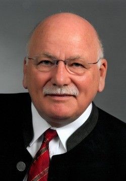 Ein kahler Mann mit Schnurrbart und Brille ist in einem schwarzen Anzug, einem weißen Hemd und einer roten Krawatte gekleidet. Er schaut direkt in die Kamera.
