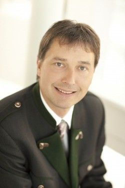Ein Mann in einem formellen Anzug und Krawatte, lächelnd und gegen einen weißen Hintergrund sitzend.