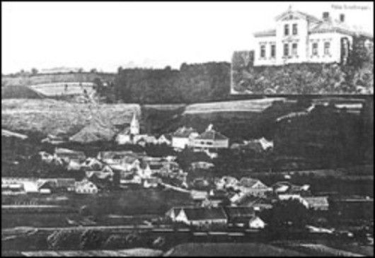 Ein schwarz-weißes Luftbild einer Stadt zeigt ein großes Gebäude auf einem Hügel und viele kleinere Häuser darunter. Bäume und Felder umgeben das Gebiet.