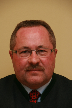 Ein Mann mit Brille und roter Punktekrawatte schaut in die Kamera. Er ist in einem schwarzen Anzug gekleidet und hat einen ernsten Ausdruck.