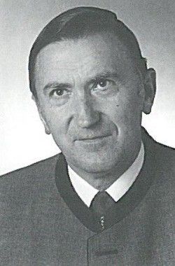 Schwarz-Weiß-Foto eines Mannes mit ernster Miene, der einen formellen Anzug und eine Krawatte trägt, mit kurzen Haaren und sichtbaren Gesichtsfalten, vor einem schlichten Hintergrund.