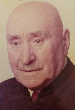 Ein älterer Mann mit kahlem Kopf und einem Schnurrbart. Er trägt einen schwarzen Anzug mit roter Krawatte. Sein Gesicht hat Falten und Muttermale.