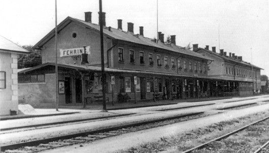 Ein Schwarz-Weiß-Bild eines alten Bahnhofs mit dem Namen 'FEHRIN' über dem Eingang, mit mehreren Fenstern, Schornsteinen und einer Plattform mit Bänken und Menschen.