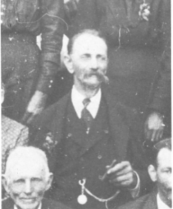 Ein Schwarz-Weiß-Foto zeigt eine Gruppe älterer Männer und Frauen, einen Mann in der Mitte mit Zigarre und Medaille, lächelnd und für ein Foto posierend.