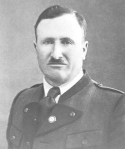Ein Schwarz-Weiß-Foto eines Mannes in Militäruniform mit einem Schnurrbart und ernster Miene, der vor einem schlichten Hintergrund steht.