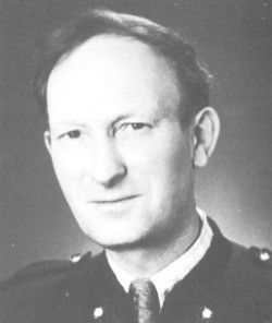 Ein Schwarz-Weiß-Foto eines Mannes in Militäruniform, der leicht lächelt und kurzes, glattes Haar hat.